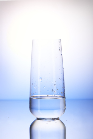One-third full drinking glass with reflection in drops of water on white and blue backgroundの写真素材