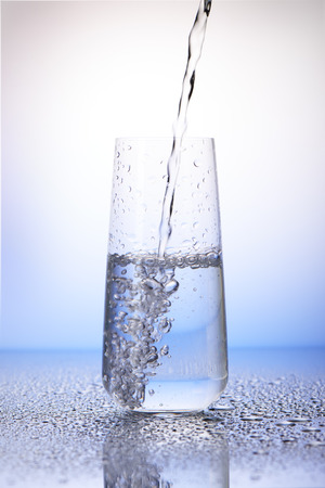 Water pouring into half-filled drinking glass with reflection in drops of water on white and blue backgroundの写真素材