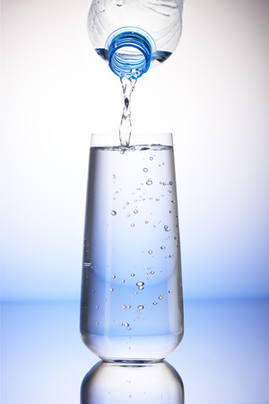 Water pouring from plastic bottle into full drinking glass with reflection on white and blue backgroundの写真素材