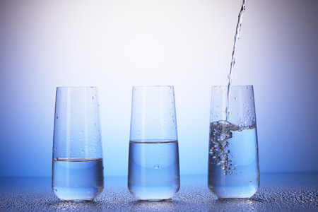 One-third full, half-filled and two-thirds full drinking glasses with reflection in drops of water on white and blue background. Water pouring into last one.の写真素材
