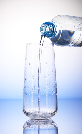 Water pouring from plastic bottle into empty drinking glass with reflection in drops of water on white and blue backgroundの写真素材