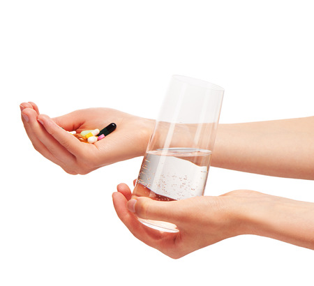 Close up of female hands holding clean transparent drinking glass with water and pile of different tablets and capsules against white background.の写真素材