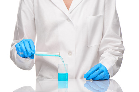 Close up of female in white gown and blue gloves pouring blue liquid from transparent white glass  test tube into measuring cup against white backgroundの写真素材