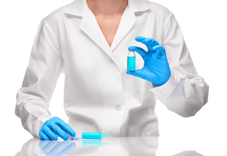 Close up of female doctor in white medical gown and blue sterilized surgical gloves holding with plastic medical syringe filled and white glass ampoule with blue drug against white backgroundの写真素材