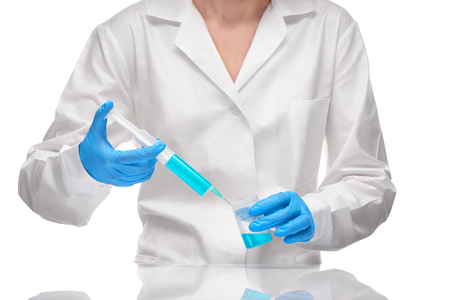 Close up of female doctor in white medical gown and blue sterilized surgical gloves holding plastic medical syringe and measuring cup with blue drug against white backgroundの写真素材