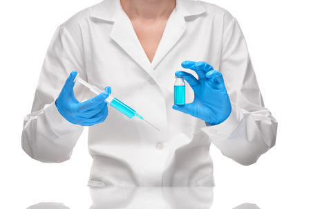 Close up of female doctor in white medical gown and blue sterilized surgical gloves holding plastic medical syringe and white glass ampoule with blue drug against white backgroundの写真素材