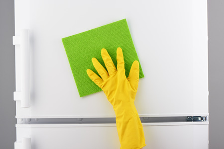 Woman's hand in yellow rubber protective glove cleaning white refrigerator with green rag on gray backgroundの写真素材