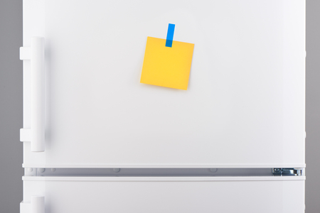 Blank yellow paper note attached with blue sticker on white refrigerator doorの写真素材