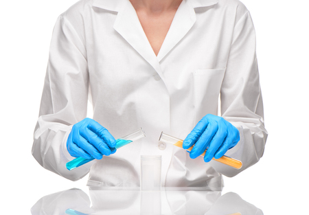Close up of female in white gown and blue gloves holding transparent white glass test tubes and with yellow sorbent and blue liquid over measuring cup against white backgroundの写真素材