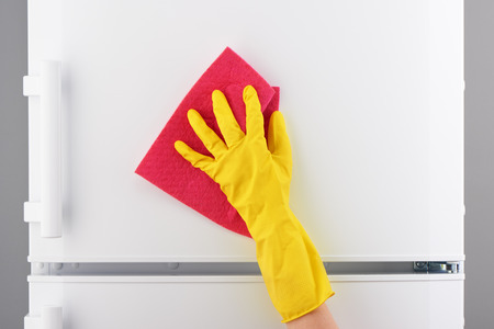 Woman's hand in yellow rubber protective glove cleaning white refrigerator with pink rag on gray backgroundの写真素材