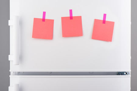Three blank pink paper notes attached with pink stickers on white refrigerator doorの写真素材