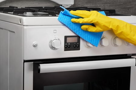 Woman's hand in yellow rubber protective glove cleaning white gas stove with blue rag on gray backgroundの写真素材