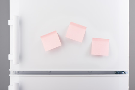 Three blank light pink sticky paper notes on white refrigerator doorの写真素材