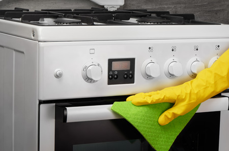Woman's hand in yellow rubber protective glove cleaning white gas stove with green rag on gray backgroundの写真素材