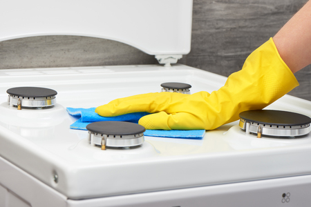 Woman's hand in yellow rubber protective glove cleaning white gas stove with blue rag on gray backgroundの写真素材