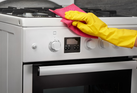 Woman's hand in yellow rubber protective glove cleaning white gas stove with pink rag on gray backgroundの写真素材