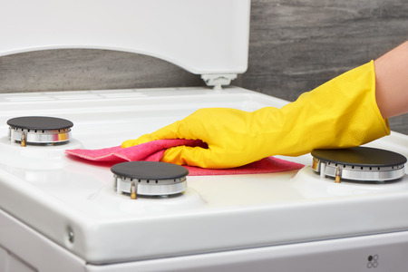 Woman's hand in yellow rubber protective glove cleaning white gas stove with pink rag on gray backgroundの写真素材