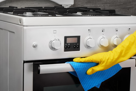 Woman's hand in yellow rubber protective glove cleaning white gas stove with blue rag on gray backgroundの写真素材