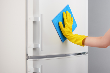 Woman's hand in yellow rubber protective glove cleaning white refrigerator with blue rag on gray backgroundの写真素材
