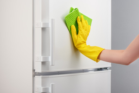 Woman's hand in yellow rubber protective glove cleaning white refrigerator with green rag on gray backgroundの写真素材