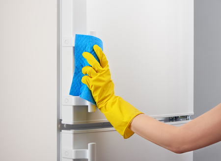 Woman's hand in yellow rubber protective glove cleaning white refrigerator with blue rag on gray backgroundの写真素材