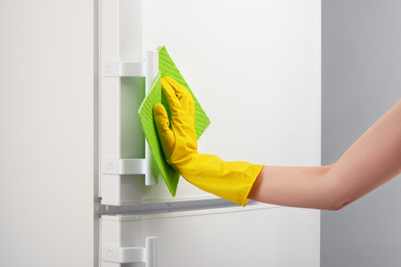 Woman's hand in yellow rubber protective glove cleaning white refrigerator with green rag on gray...