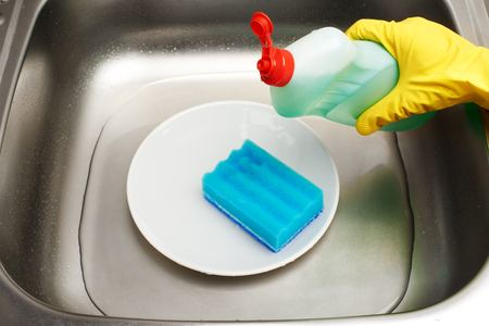 Close up of kitchen sink with water, clean white plate, blue cleaning sponge and bottle of liquid detergent in female hand in yellow protective rubber gloveの写真素材