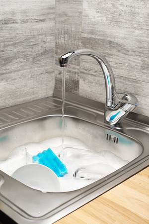 Close up of kitchen sink with running tap water full of foam, tableware and blue cleaning spongeの写真素材