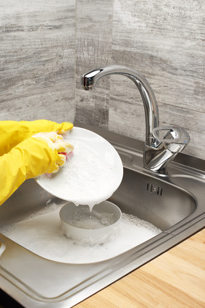 Close up of female hands in yellow protective rubber gloves washing white plate with pink cleaning sponge against kitchen sink full of foam and tablewareの写真素材