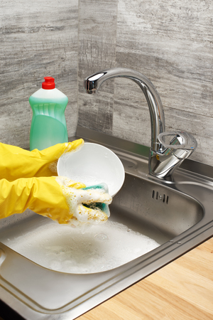 Close up of female hands in yellow protective rubber gloves washing tableware with green cleaning sponge and liquid detergent in bottle against kitchen sink full of foamの写真素材