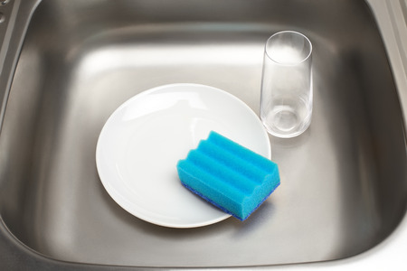 Close up of kitchen sink with clean white plate, blue cleaning sponge and drinking glassの写真素材