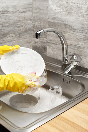 Close up of female hands in yellow protective rubber gloves washing white plate with pink cleaning sponge against kitchen sink full of foam and tablewareの写真素材