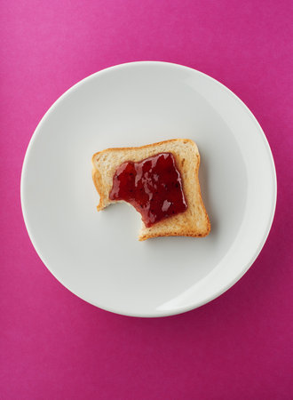Crispy slice of toast with strawberry jam and one bite missing on white plate and bright pink backgroundの写真素材