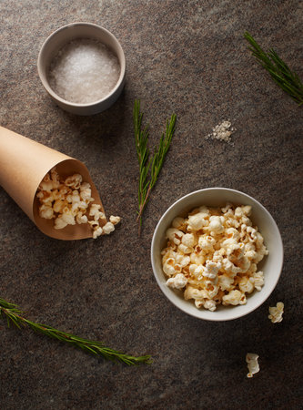 White porcelain bowl and brown paper cone full of popcorn with salt and green sprigs of rosemary on marble countertopの写真素材