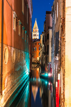 A narrow canal in Venice, Italy, at night, with a boat and a tall bell tower of St Marks Campanile in the distanceの写真素材