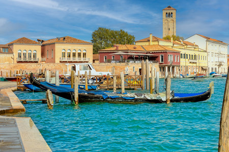 Traditional Venetian waterfront scene on Murano island with gondolas, colorful buildings, and a historic bell tower under sunny skies.の写真素材