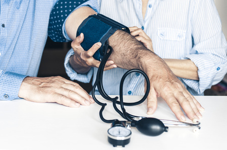The therapist female hands with a classic tonometer measuring blood pressure of an man in home room on white tableの写真素材