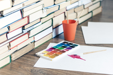 Watercolor paint, brushes well used and sheet of paper on wooden table with stack of books backgroundの写真素材