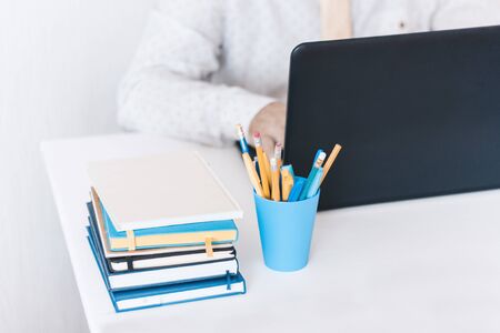 Close-up of hands middle-aged man in white shirt and yellow tie typing on keyboard laptop computer, plastic blue holder with pencils and pens, stack of books, notebooks, smartphone, glasses on white table, education office concept backgroundの写真素材