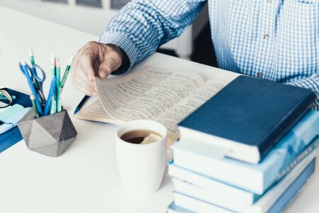 Middle aged man in blue shirt reading book on modern stylish work place with office supplies,...