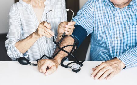 The therapist male hands in blue shirt with a classic tonometer measuring blood pressure of a man in home room on white tableの写真素材