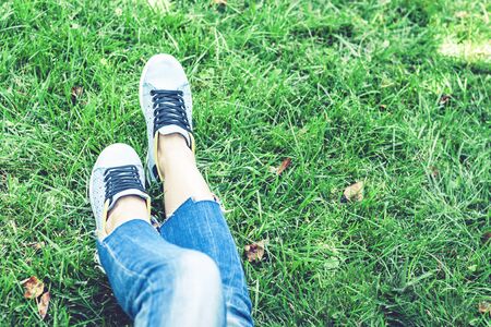 Young woman legs in sport shoes sneakers of blue suede, sitting on the grass lawn in parkの写真素材