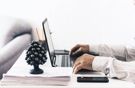 Close-up of hands middle-aged man in white shirt typing on keyboard laptop computer, holder with pencils and pens, stack of books, notebooks, smartphone, glasses on white table, education office concept backgroundの写真素材