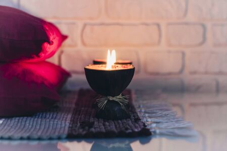 Burning spa aroma candle in coconut shell on a knitted multicolored rug and red pillows opposite the...