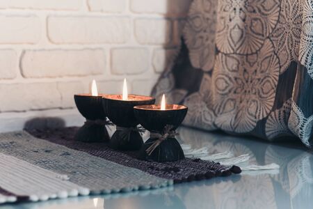 Burning spa aroma candle in coconut shell on a knitted multicolored rug and gray curtains opposite...