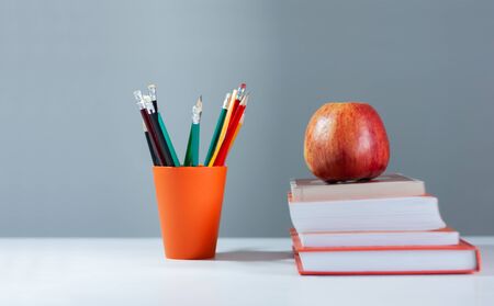 Orange pencil holder, stack of books on white table with red apple. Books stacking. Back to school...