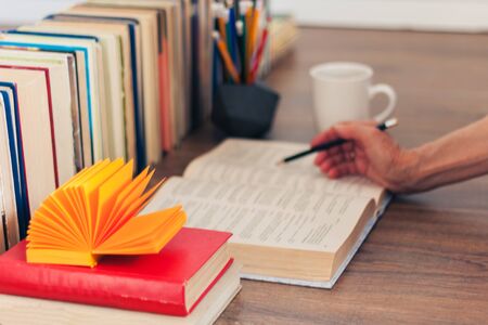 Hand of teacher, open textbook, notebook, glasses, pencils in holder, cup of tea and stack of old book on wooden table, education concept background, many books piles with copy space for textの写真素材