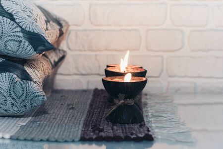 Burning spa aroma candle in coconut shell on a knitted multicolored rug and gray pillows opposite the white wall, cozy home interiorの写真素材