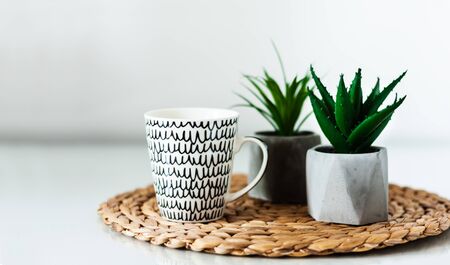 Cozy home interior decor: white and black cup and ornamental plants in pots on a wicker stand on a white table in the room. The quarantine concept of stay homeの写真素材