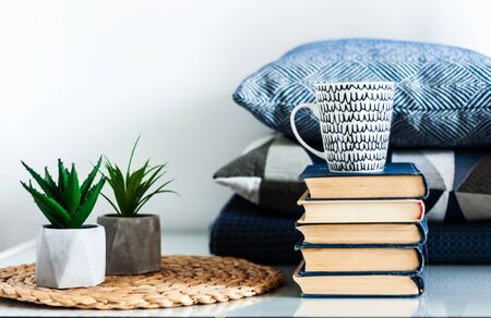 Cozy home interior decor: white and black cup, stack of books, plants in pots on a wicker stand, blue pillows on a white table in the room. Distance home education. Quarantine concept of stay homeの写真素材
